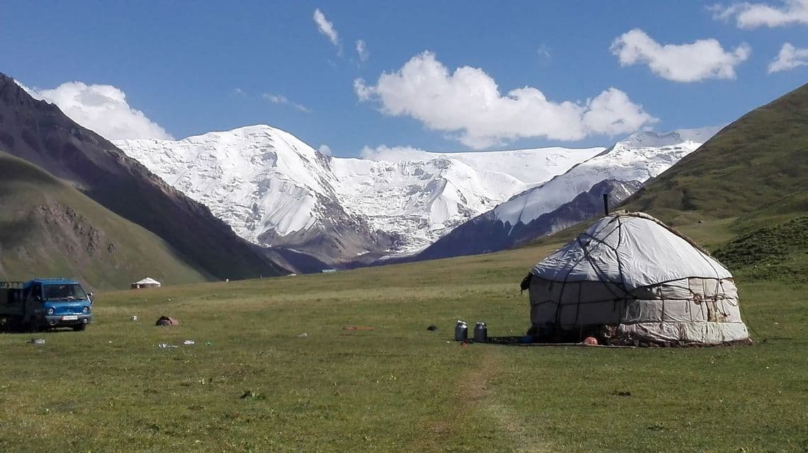 Una yurta tradizionale e un camion blu si trovano in una vasta valle verde di fronte a una grande catena montuosa innevata sotto un cielo blu.
