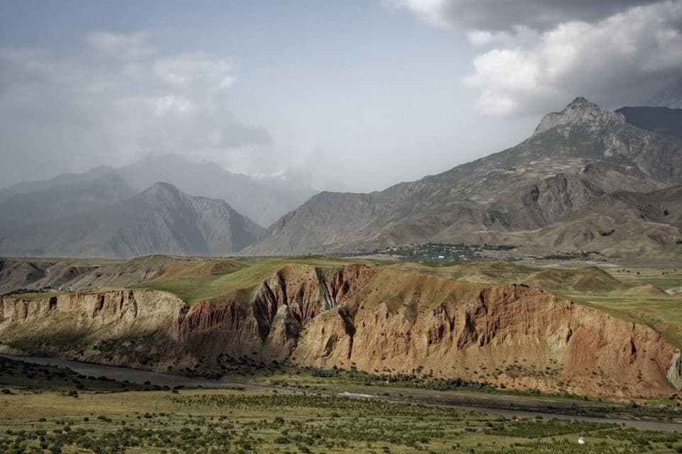 Ampi canyon rossastri fiancheggiano un fiume in una vasta valle di montagna, con un villaggio distante annidato tra verdi colline sotto un cielo nuvoloso.