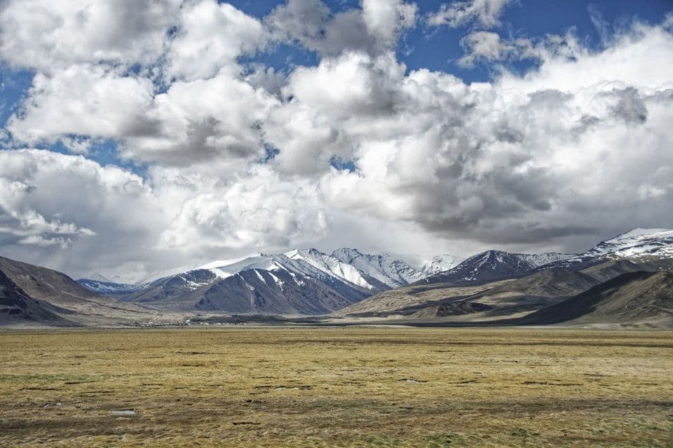 Una vasta pianura erbosa si estende verso una catena di montagne innevate sotto un cielo pieno di soffici nuvole bianche.