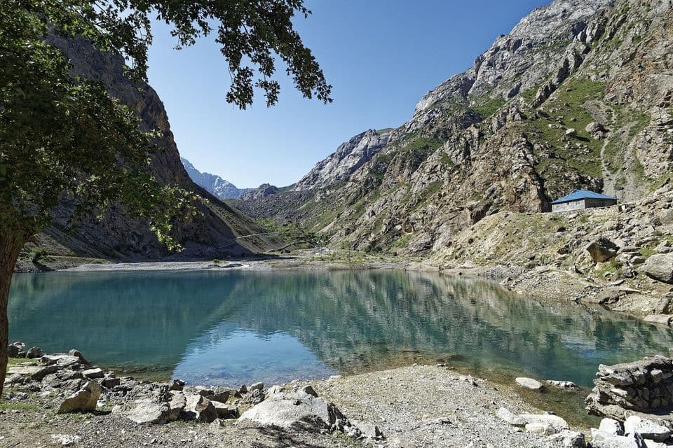 Un lago calmo e turchese si adagia in una valle montuosa rocciosa, riflettendo le cime e una piccola casa sulla riva.