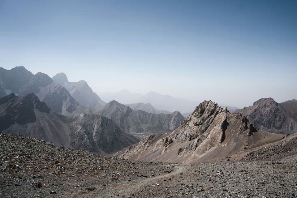Un sentiero escursionistico si snoda giù per un ripido versante montuoso roccioso, affacciandosi su una valle con un piccolo lago turchese e cime sfocate in lontananza.
