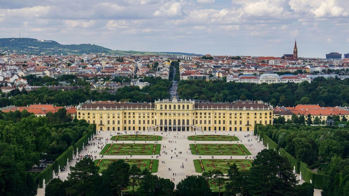 Una vista aerea di un grande palazzo giallo con i suoi ampi giardini formali, con uno skyline cittadino visibile sullo sfondo.