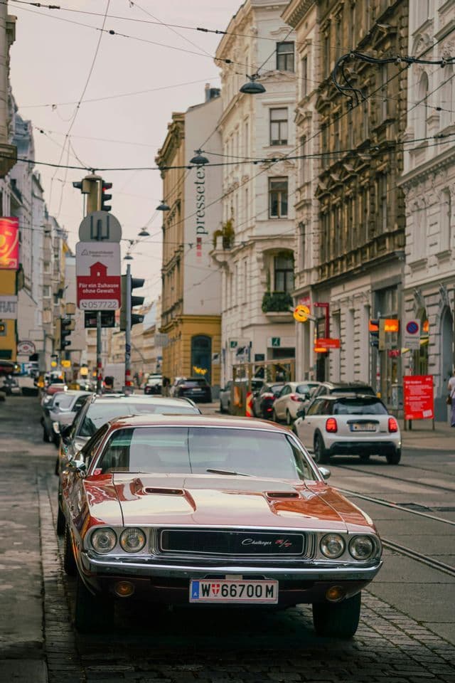 Una Dodge Challenger rossa d'epoca parcheggiata su una strada acciottolata di città, con classici edifici europei sullo sfondo.