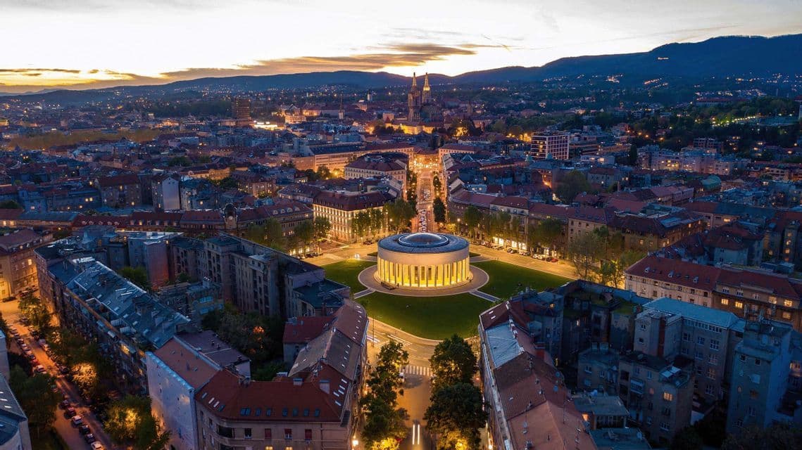 Eine Luftaufnahme einer Stadt in der Abenddämmerung, mit einem beleuchteten kreisförmigen Gebäude auf einem zentralen Platz und Bergen am Horizont.