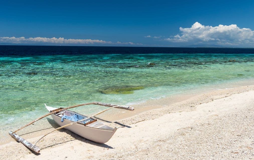 Una canoa con balancín descansa en una playa de arena blanca junto a agua turquesa cristalina bajo un cielo azul.