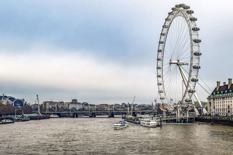 Una grande ruota panoramica si erge sulla riva di un ampio fiume, con barche sull'acqua e uno skyline cittadino sullo sfondo.
