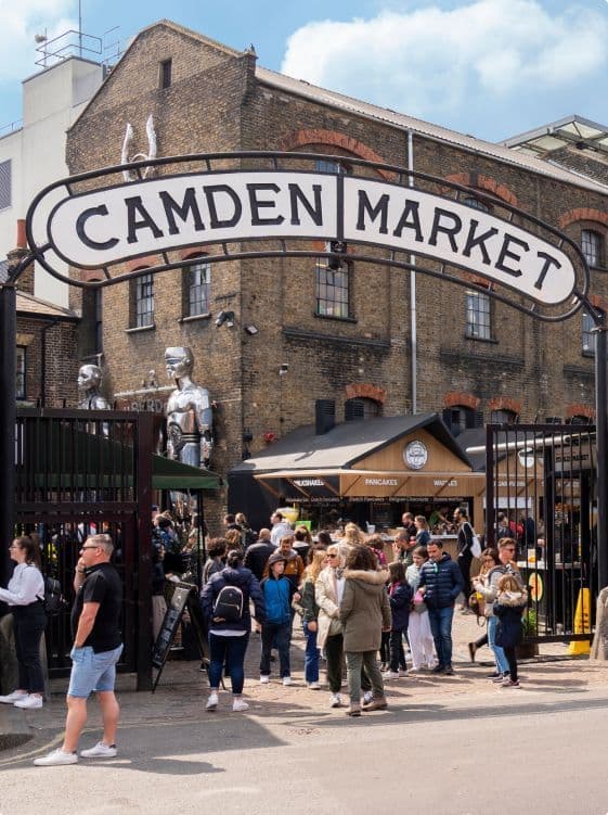 Una folla di persone si trova all'ingresso del Camden Market, sotto un grande cartello ad arco, con due statue di robot argentati vicino al cancello.