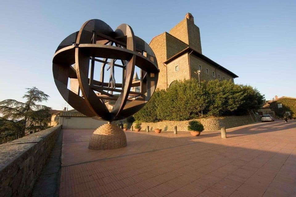 Una grande scultura in metallo di una sfera armillare su una base di mattoni si erge su una piazza pavimentata di fronte a un edificio in pietra sotto un cielo sereno.