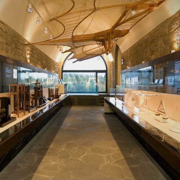 Un grande modello in legno di una macchina volante storica è appeso al soffitto di una galleria museale con pareti in pietra e teche di vetro.