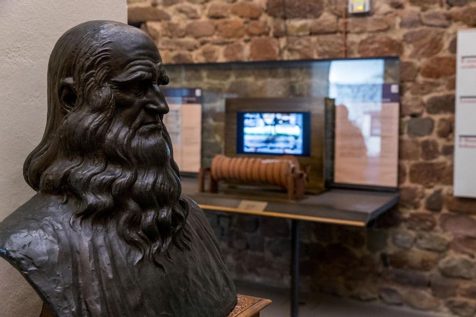 Un busto in bronzo di Leonardo da Vinci, mostrato di profilo all'interno di un museo, con un'esposizione delle sue invenzioni e un muro di pietra dietro.