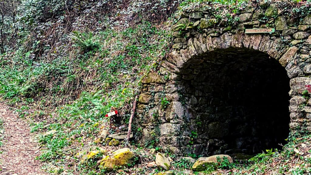 L'ingresso ad arco di un vecchio tunnel in pietra incastonato in una collina ricoperta di vegetazione verde, con un segnavia nelle vicinanze.