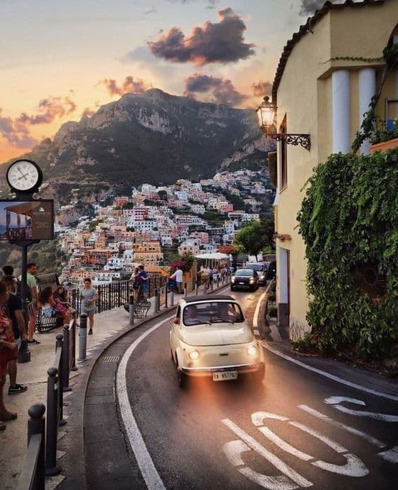 Un coche clásico con las luces encendidas sube por una carretera sinuosa a través de un pueblo en el acantilado, con montañas al fondo al atardecer.