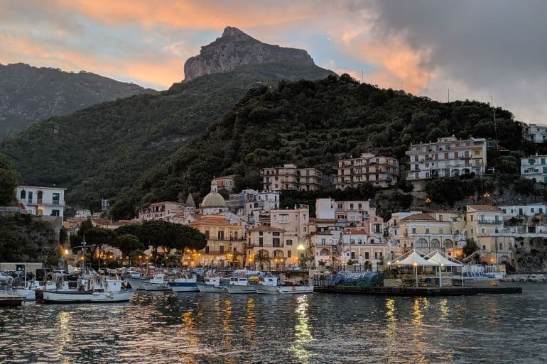 Un pueblo costero con un puerto lleno de barcos anidado al pie de una montaña verde al atardecer.