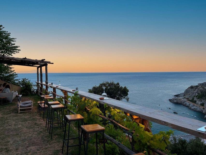 Un bar al aire libre con una larga barra de madera y taburetes en una terraza con vistas al mar al atardecer.