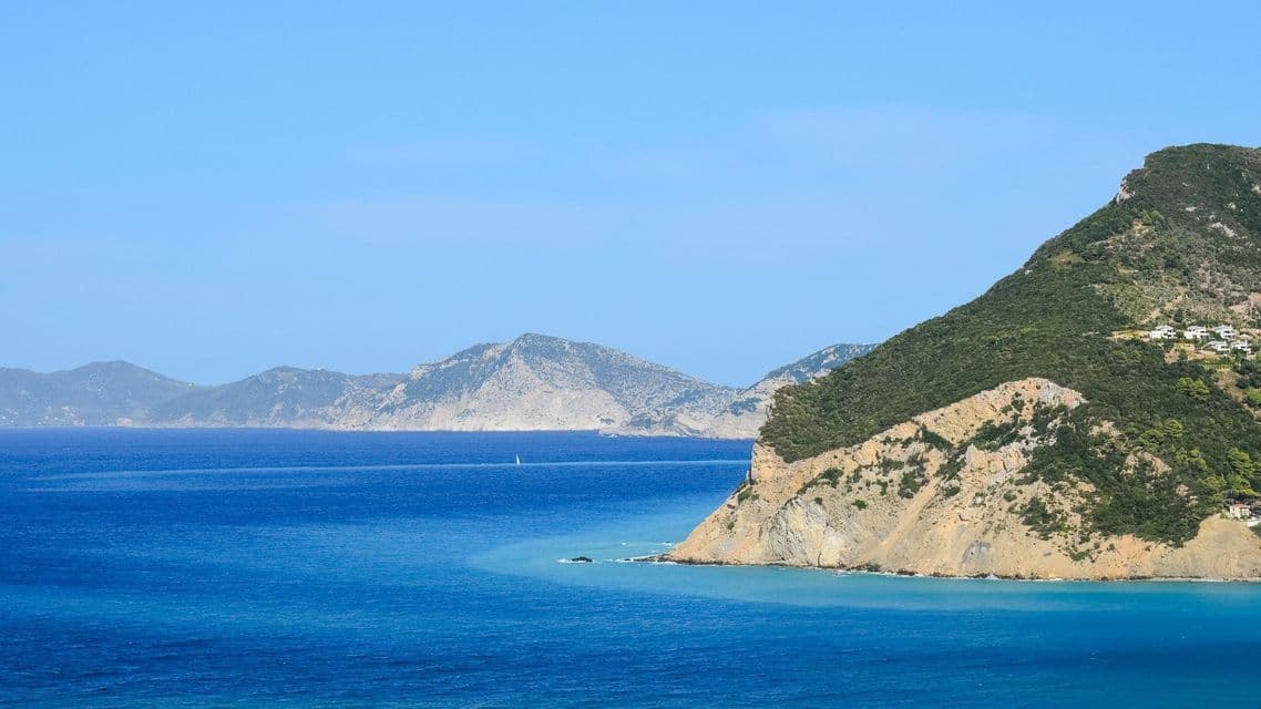 Una scogliera verde e rocciosa si protende nel mare blu profondo, con montagne sfocate all'orizzonte sotto un cielo limpido.
