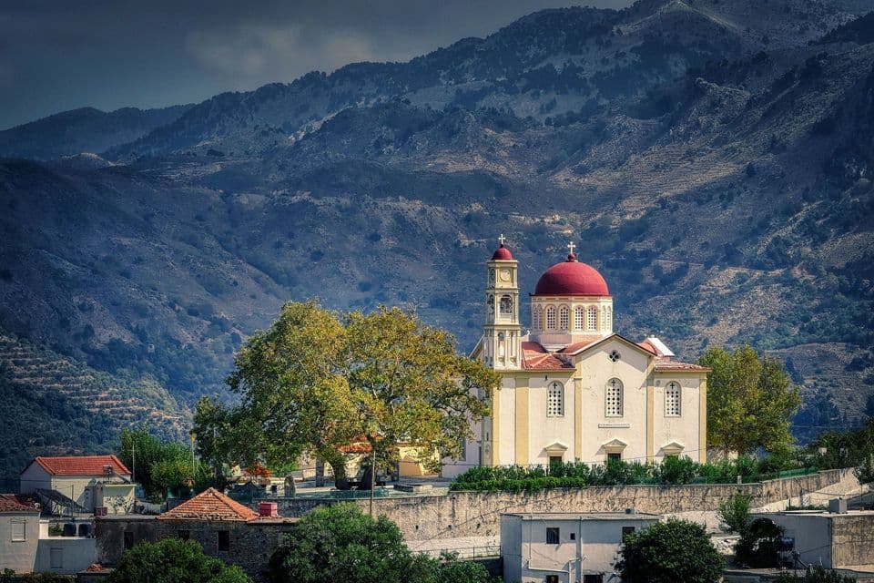 Una chiesa bianca con una cupola rossa e un campanile si erge su una collina terrazzata di fronte a uno sfondo di montagne aspre