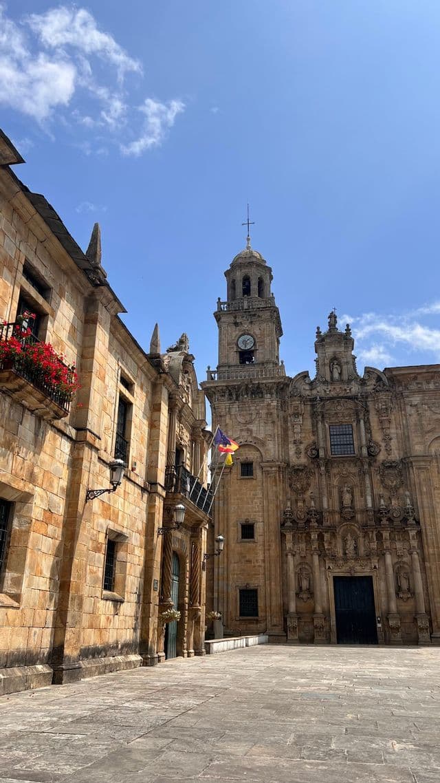 Edifici in pietra decorati con una prominente torre dell'orologio si ergono in una piazza pavimentata sotto un cielo blu con nuvole sparse.