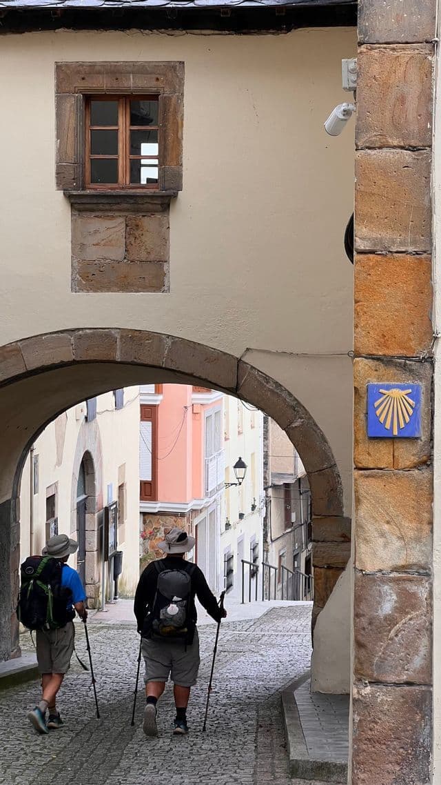Due pellegrini WeRoad con zaini e bastoni da trekking camminano sotto un arco in pietra su una strada acciottolata, contrassegnata dal simbolo della conchiglia di capasanta.