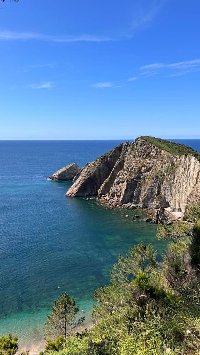 Una scogliera rocciosa e dalla cima verde si staglia in un oceano calmo e blu in una giornata di sole, con una piccola spiaggia e bagnanti visibili al di sotto.