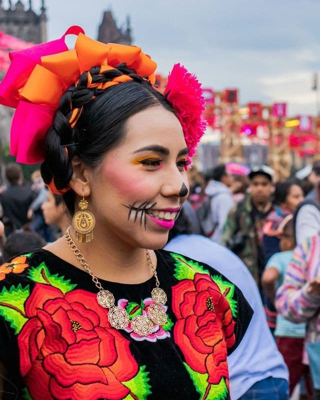 Una donna con trucco Calavera colorato, un abito floreale ricamato e un grande copricapo a fiocco sorride durante un festival.
