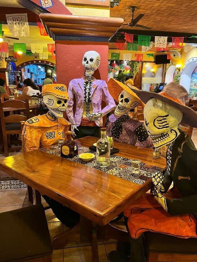 Quattro figure colorate di scheletri del Giorno dei Morti siedono a un tavolo di legno di un ristorante con bottiglie di tequila.
