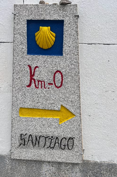 Ein Granitwegweiser für den Jakobsweg, der eine gelbe Jakobsmuschel, „Km-0“ und einen Pfeil in Richtung Santiago zeigt.