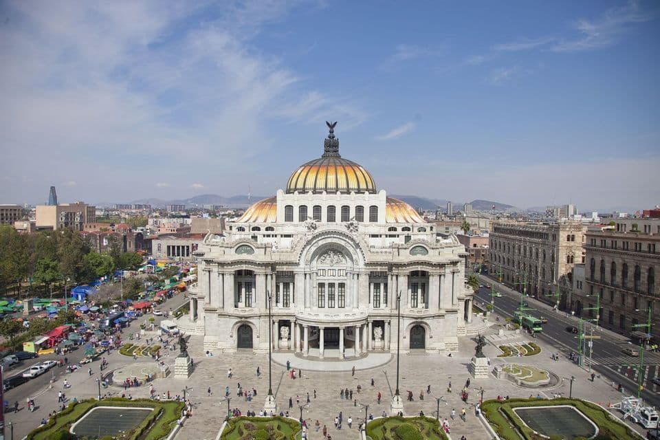 Un edificio bianco e sontuoso con una cupola dorata, posizionato al centro di una vivace piazza cittadina sotto un cielo azzurro splendente, con un panorama urbano sullo sfondo.
