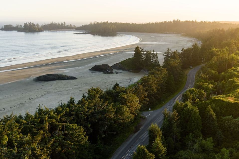 Vue aérienne d'une route sinueuse traversant une forêt de conifères le long d'une large plage de sable au coucher du soleil.