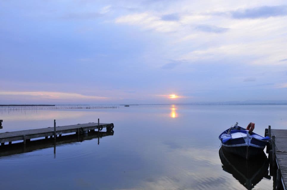 Un barco azul amarrado a un embarcadero de madera en aguas tranquilas mientras el sol se pone en el horizonte, reflejándose en el lago.