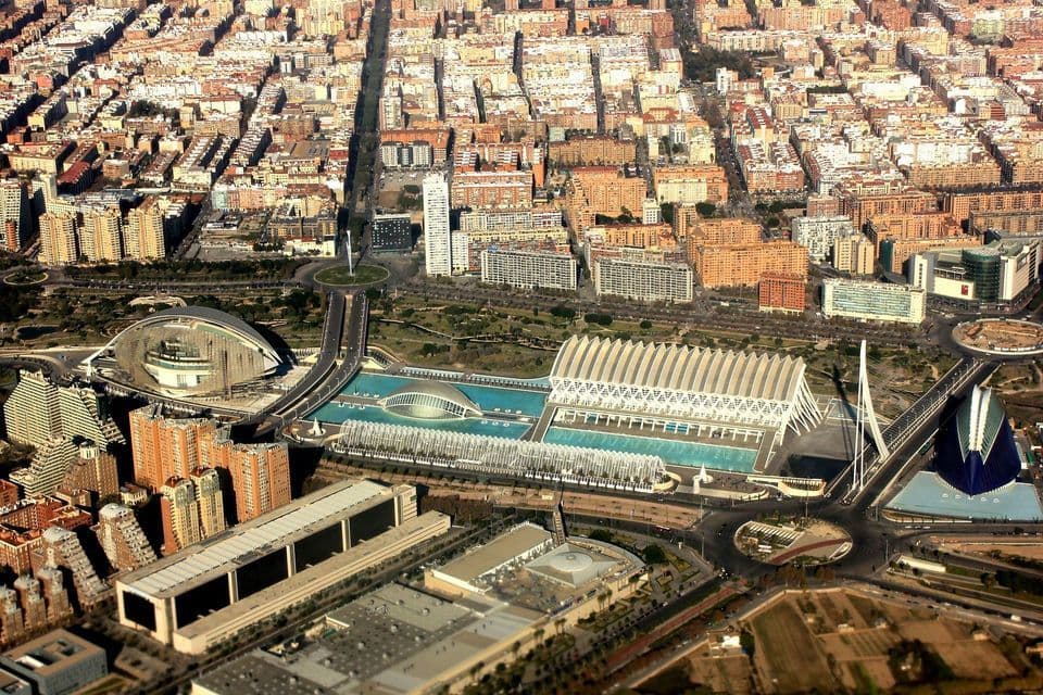 Una vista aérea de un complejo arquitectónico futurista con edificios blancos y piscinas azules, ubicado en una ciudad grande y extensa.