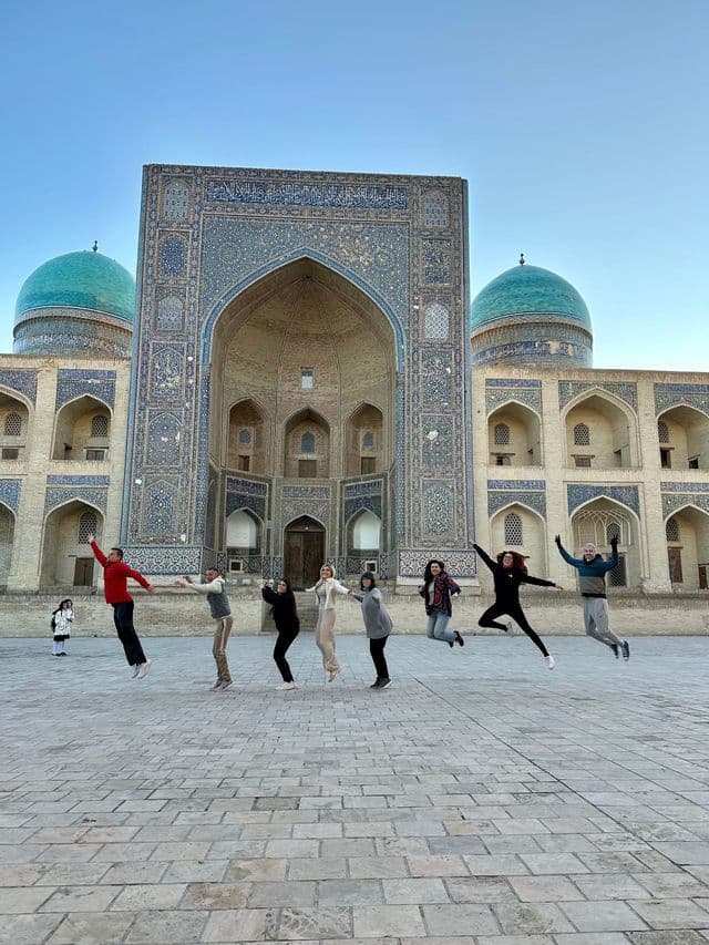 A WeRoad group trip jumps in the air in a stone courtyard in front of an ornate building with turquoise domes.