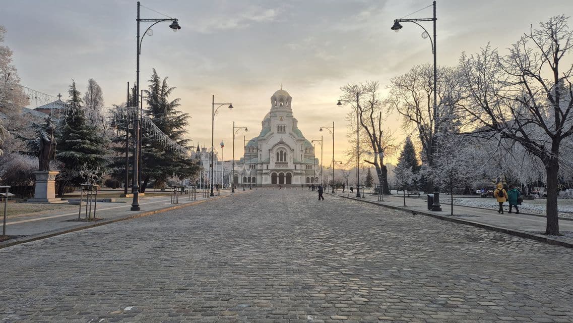 Una calle ancha y adoquinada conduce hacia una gran catedral con cúpula, flanqueada por árboles cubiertos de escarcha y farolas al amanecer.
