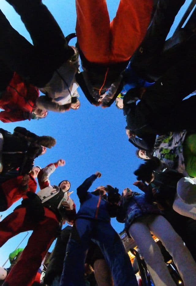 A WeRoad group trip in winter clothes huddles in a circle, seen from a low angle against a clear blue sky.