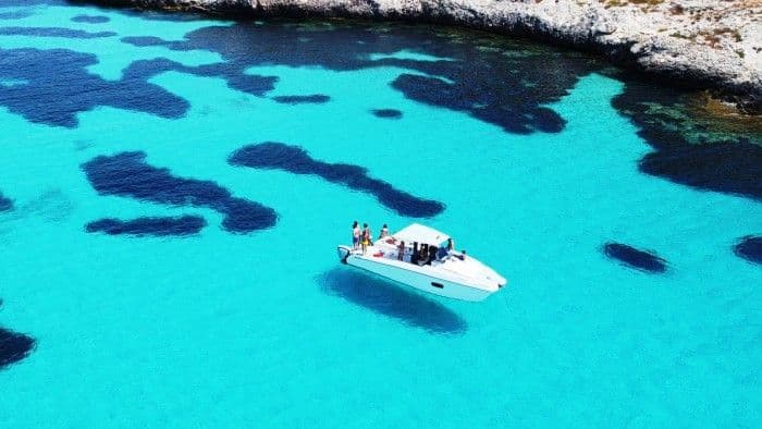 Veduta aerea di un viaggio di gruppo WeRoad su un motoscafo bianco che naviga in acque limpide e turchesi vicino a una costa rocciosa.