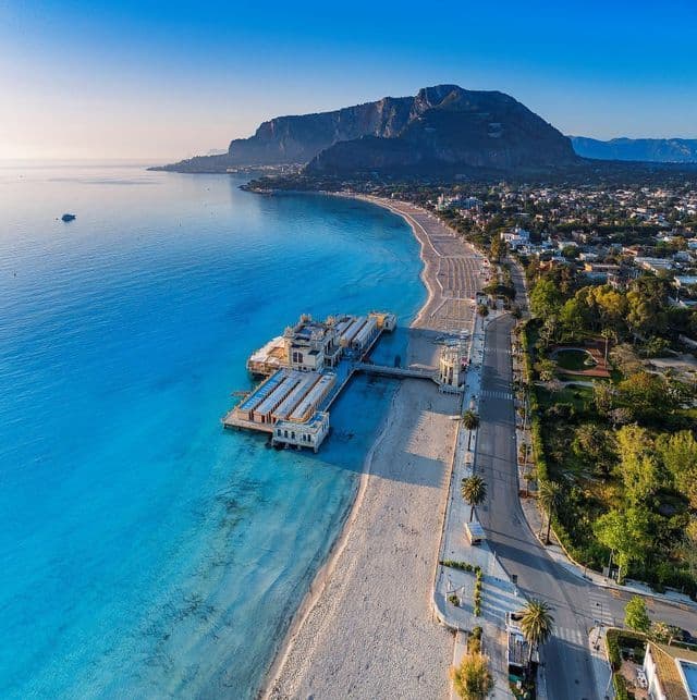 Una vista aerea di una lunga spiaggia sabbiosa con un molo ornato, acqua turchese e una grande montagna sullo sfondo.