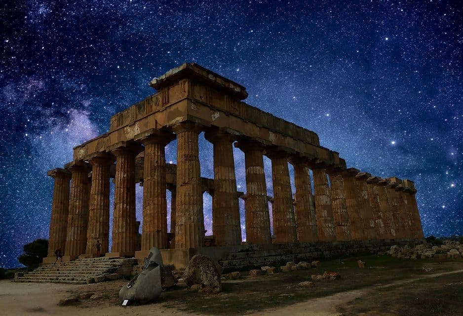 Un antico tempio di pietra con grandi colonne consumate dal tempo è illuminato contro un cielo buio e stellato.