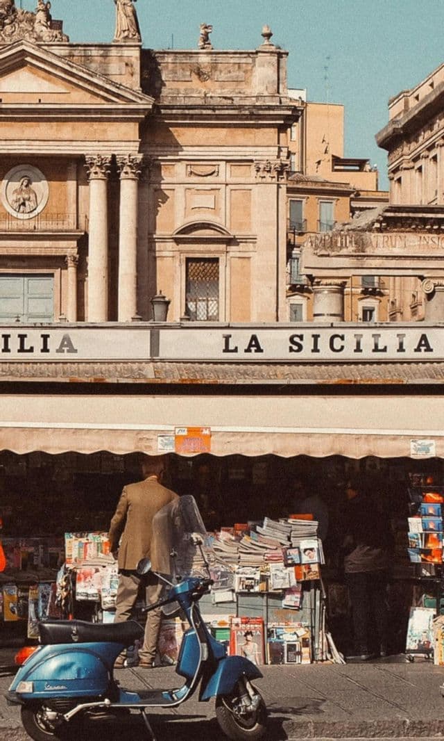 Uno scooter blu d'epoca è parcheggiato su una strada di fronte a un'edicola con un'insegna 'La Sicilia' sotto un edificio storico.
