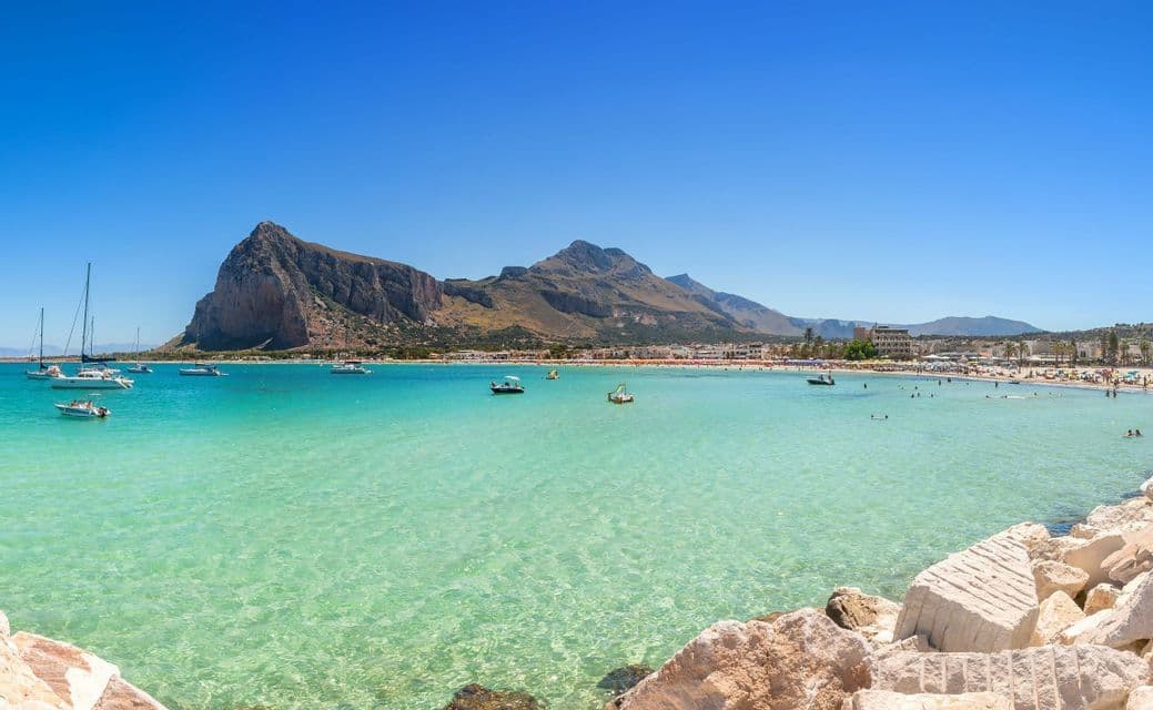 Una vista di spiaggia soleggiata con barche sull'acqua turchese, una spiaggia affollata e una grande montagna che si staglia contro un cielo azzurro.