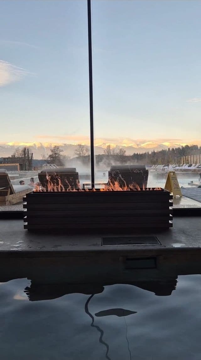 A fire pit burning beside an outdoor thermal pool with people bathing against a backdrop of snowy mountains.