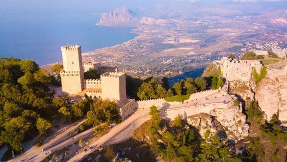 Una vista aerea di un castello di pietra con torri su una collina verdeggiante che domina una costa, il mare e una montagna distante.