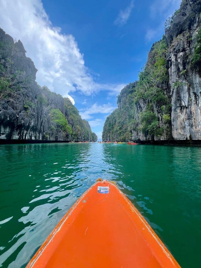 Un viaggio di gruppo WeRoad in kayak attraverso una laguna con acqua verde smeraldo, visto dalla prua di una barca arancione tra alte scogliere.