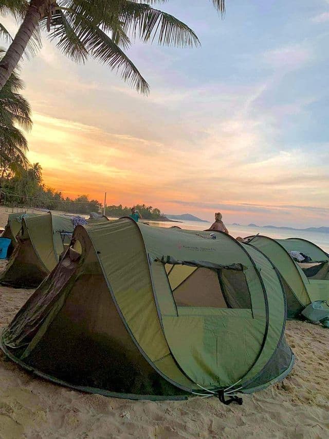 Un viaggio di gruppo WeRoad in campeggio con tende verdi su una spiaggia sabbiosa con palme all'alba.