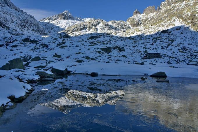 Una cordillera nevada con una cabaña de piedra reflejada en el agua cristalina de un lago alpino parcialmente congelado.
