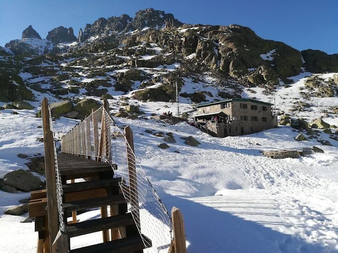 Un puente de madera con una barandilla de red conduce por una ladera de montaña nevada hasta un refugio de piedra.