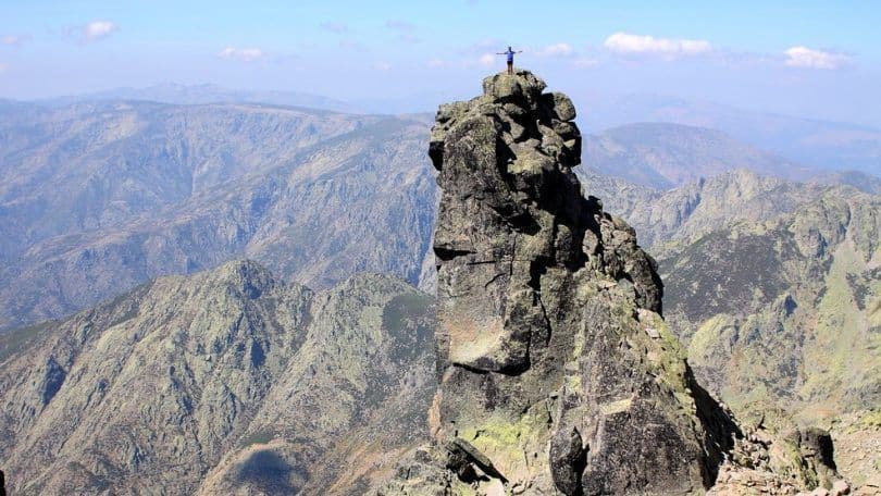Una persona se alza con los brazos extendidos en la cima de un escarpado pico rocoso, contemplando una vasta cordillera.