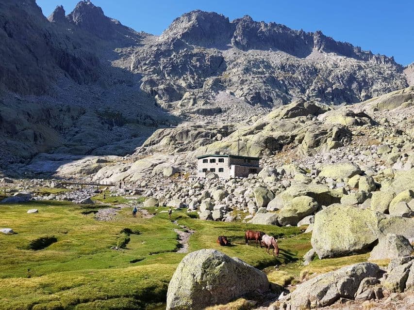 Varios caballos pastan en un prado verde cerca de un edificio de piedra al pie de una escarpada cordillera rocosa.