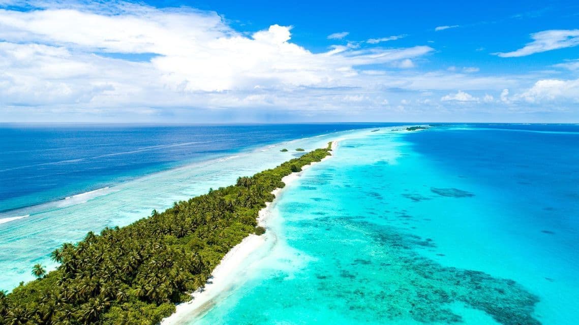 Vue aérienne d'une île tropicale étroite avec palmiers et plage de sable blanc, séparant des eaux turquoise d'un océan d'un bleu profond.