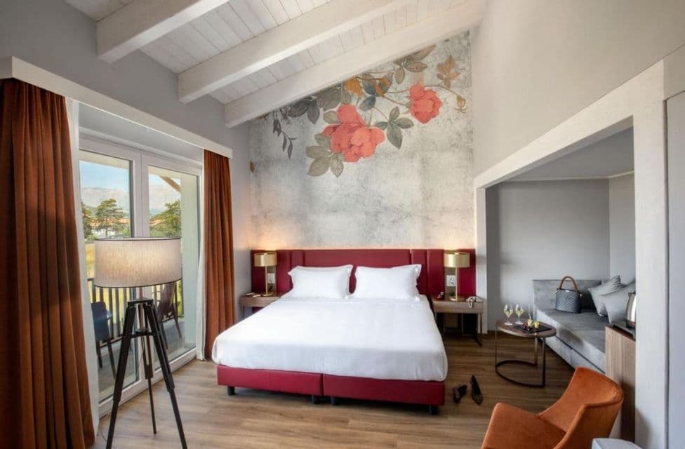 Una suite d'hotel con letto rosso, parete floreale d'accento, area salotto e balcone con vista esterna.