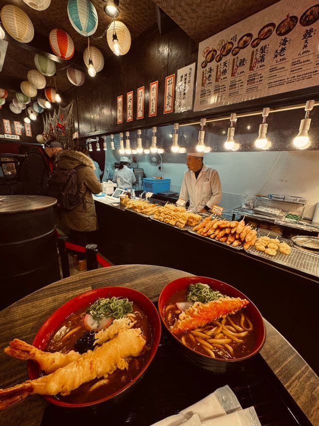 Due ciotole di udon tempura di gamberi su un tavolo in un vivace ristorante giapponese decorato con lanterne di carta.