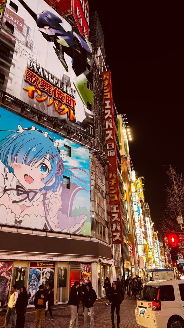 Alti edifici coperti da grandi cartelloni anime e insegne al neon illuminano una trafficata via della città giapponese di notte.